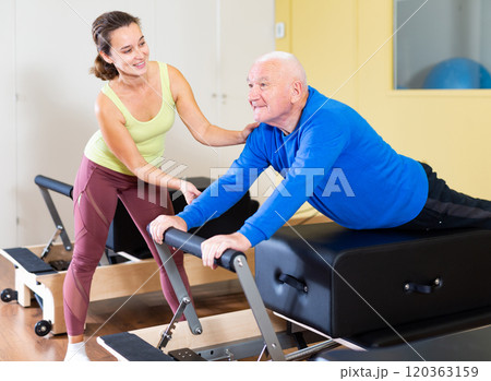 Instructor of pilates helping elderly man workout. Two people working in pilates studio, woman assistant supporting and correcting old man patient beginner. 120363159