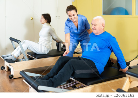 Medical professional helps and monitors the difficulty of Pilates exercise for an mature man 120363162