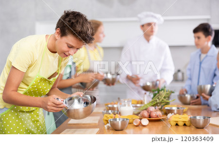 Cooking course - portrait of teenage boy in an apron who is learning to beat eggs to make pancakes at cooking class 120363409