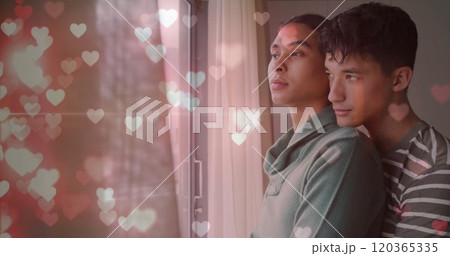 Image of glittering hearts over happy diverse gay male couple looking out of window, embracing 120365335