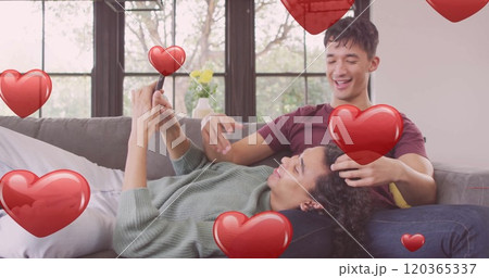 Image of red hearts over happy diverse gay male couple relaxing on couch using smartphone 120365337