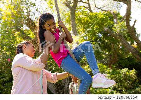Mother helping daughter swing on rope in backyard, enjoying family time outdoors 120366154