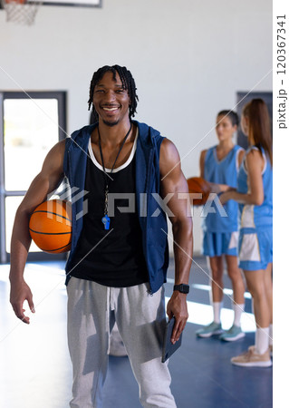 male african American basketball coach holding ball and whistle, smiling in gym with female players male african American basketball coach holding ball and whistle, smiling in gym with female players 120367341