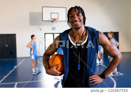 male african American basketball coach holding ball, smiling in gym with female players practicing male african American basketball coach holding ball, smiling in gym with female players practicing 120367350