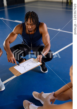 Male coach explaining basketball strategy on clipboard to female players in gym 120367420