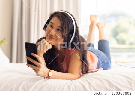 Listening to music with headphones, Asian woman relaxing and holding smartphone on bed 120367802