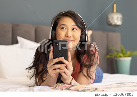 Listening to music with headphones, Asian woman holding smartphone and smiling on bed 120367803