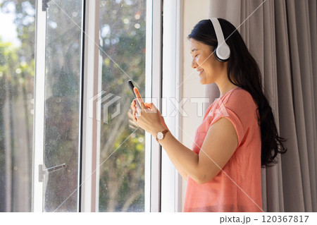 Wearing headphones, Asian woman using smartphone and smiling while standing by window 120367817