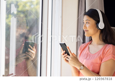 Listening to music on headphones, Asian woman holding smartphone and looking outside, copy space Listening to music on headphones, Asian woman holding smartphone and looking outside, copy space 120367818