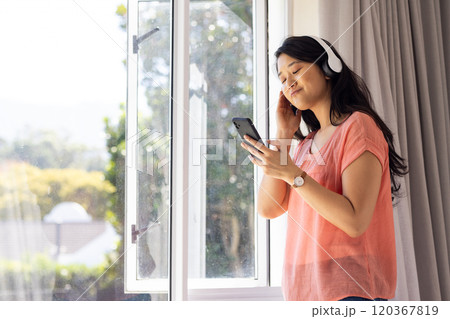 Listening to music on headphones, Asian woman enjoying smartphone by window, copy space 120367819
