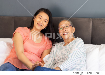 Smiling Asian grandmother and granddaughter sitting together on bed, enjoying time Smiling Asian grandmother and granddaughter sitting together on bed, enjoying time 120367838