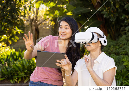 Using VR headset, asian grandmother exploring virtual reality with assistance outdoors 120368735