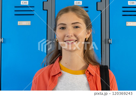 Smiling teenage girl standing in front of blue lockers at school, carrying backpack 120369243