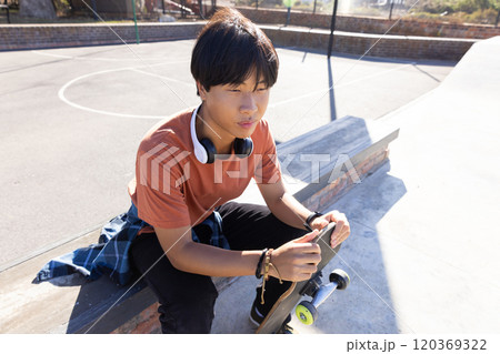 Outdoors, Sitting on bench, asian teenage boy holding skateboard and wearing headphones at skatepark 120369322