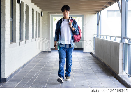 Walking in school hallway, asian teenage boy carrying backpack and books, looking ahead 120369345