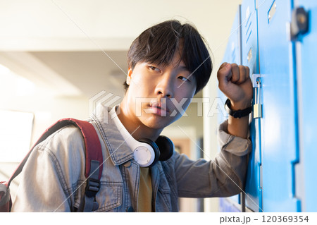 Opening locker, asian teenage boy with headphones looking away in school hallway 120369354