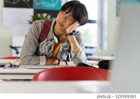 In school, asian teenage boy sitting at desk looking bored with headphones around neck In school, asian teenage boy sitting at desk looking bored with headphones around neck 120369361
