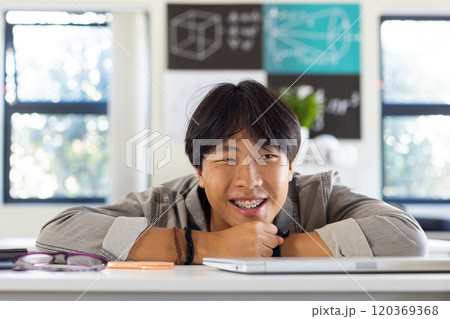 Smiling asian teenage boy with braces leaning on desk in school classroom, feeling relaxed Smiling asian teenage boy with braces leaning on desk in school classroom, feeling relaxed 120369368