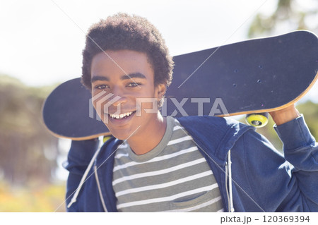 Smiling african american teenage boy holding skateboard, enjoying outdoor hangout on sunny day Smiling african american teenage boy holding skateboard, enjoying outdoor hangout on sunny day 120369394