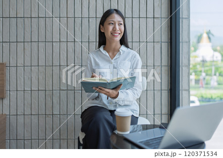 Young woman sitting in a modern cafe, smiling while taking notes in a notebook, with a laptop and coffee cup on the table, enjoying a productive study session. 120371534