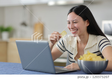 Happy asian woman using laptop eating potato chips Happy asian woman using laptop eating potato chips 120373207