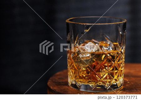 A whiskey glass resting on a wooden table, set against a dark background 120373771