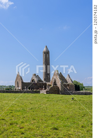 Kilmacduagh Abbey ruins in County Galway, Ireland, showcase a tall round tower and the remains of an ancient stone church. This vertical photo captures the historic site under a clear blue sky Kilmacduagh Abbey ruins in County Galway, Ireland, showcase a tall round tower and the remains of an ancient stone church. This vertical photo captures the historic site under a clear blue sky 120374182