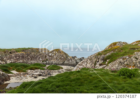 Coastal landscape of Coral Strand Beach, Connemara, Ireland, showcasing rugged rocks and lush green hills leading down to a fog-shrouded sea 120374187