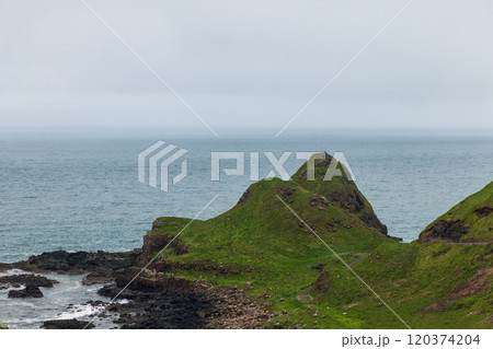 Rugged Irish shoreline where green hills meet Atlantic Ocean beneath an overcast grey sky 120374204