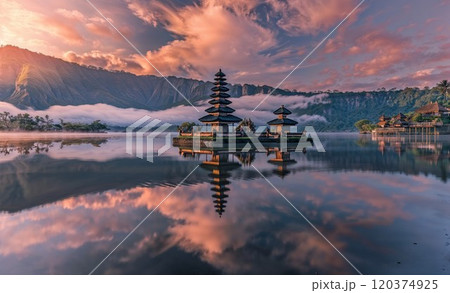 A serene Balinese temple reflecting in calm lake waters under a breathtaking sunrise. Misty mountains and soft clouds enhance the spiritual and tranquil atmosphere 120374925