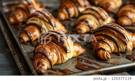 photo shows freshly baked croissants drizzled with chocolate and dusted with powdered sugar on a baking tray. The croissants have a golden-brown, flaky crust, creating an inviting and delicious scene 120377114