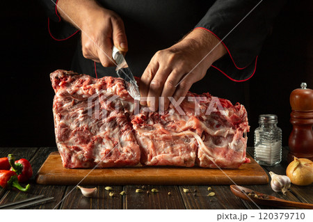 The process of grilling on the kitchen table. A knife in the hands of a butcher before cutting meat. A large piece of ribs on a cutting board and aromatic spices 120379710