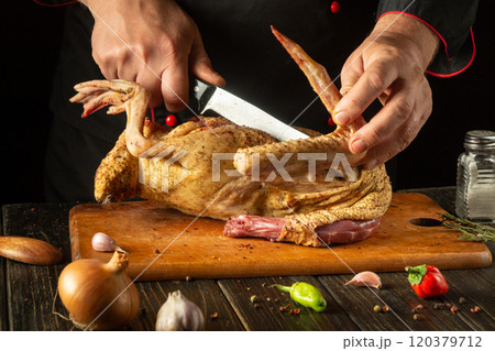 A skilled chef adeptly carves a whole chicken with a large knife, surrounded by fresh ingredients. The warm kitchen atmosphere highlights the culinary artistry in preparation 120379712