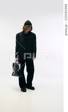 Fashion-forward look. Full-length portrait of stylish young guy in beanie hat, total black costume, glasses and leather bag, posing against white studio background Fashion-forward look. Full-length portrait of stylish young guy in beanie hat, total black costume, glasses and leather bag, posing against white studio background 120381666