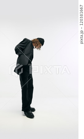 Side view full-length image of young man wearing total black outfit, blazer, pants and beanie hat, posing against white studio background Side view full-length image of young man wearing total black outfit, blazer, pants and beanie hat, posing against white studio background 120381667