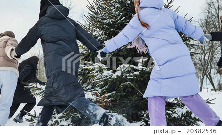 Schoolgirls joyfully running around a christmas tree, holding hands in a snowy winter park 120382536