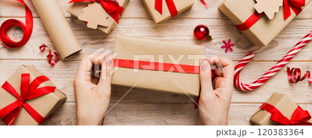 Woman is packing surprise gift for Christmas holidays, sitting at workplace with craft tools, top view. Woman wrapping Christmas gift at colored table 120383364