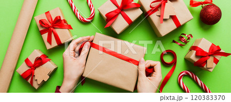 Woman is packing surprise gift for Christmas holidays, sitting at workplace with craft tools, top view. Woman wrapping Christmas gift at colored table 120383370