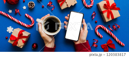 Above view of female hand holding smart phone with hot cup of coffee. Christmas decoration on colored background 120383817
