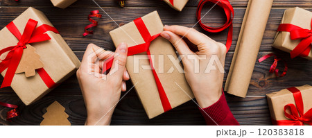 Woman is packing surprise gift for Christmas holidays, sitting at workplace with craft tools, top view. Woman wrapping Christmas gift at colored table 120383818