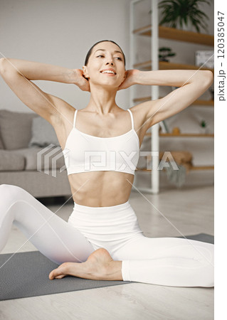 Attractive young woman doing yoga stretching at home. Brunette woman wearing white leggins and top. Self-isolation is beneficial, entertainment and education on the Internet. 120385047