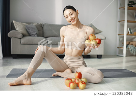 Slim and beautiful woman wearing beige leggins and top and sitting on yoga mat. Young girl with fit body posing at home with fresh apples. Sport, fitness, diet, nutrition and health care concept. Slim and beautiful woman wearing beige leggins and top and sitting on yoga mat. Young girl with fit body posing at home with fresh apples. Sport, fitness, diet, nutrition and health care concept. 120385048