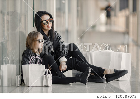 Mother and daughter on shopping. Young pretty clients standing in front of glasses wall holding shopping paper bags. Brunette woman with her daughter wearing black clothes. Mother and daughter on shopping. Young pretty clients standing in front of glasses wall holding shopping paper bags. Brunette woman with her daughter wearing black clothes. 120385075