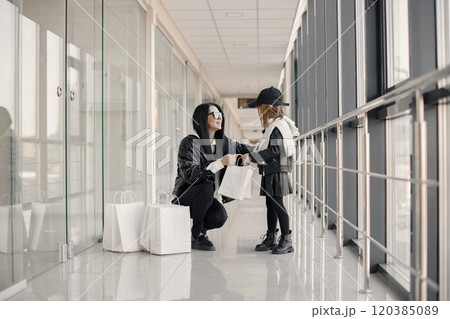 Mother and daughter on shopping. Young pretty clients standing in front of glasses wall holding shopping paper bags. Brunette woman with her daughter wearing black clothes. 120385089