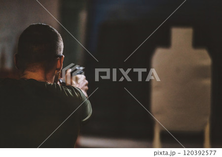 Man aiming a firearm toward a target at what appears to be an indoor shooting range. man is seen from behind, focusing on the target in front of them. 120392577