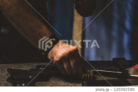 Man handling ammunition. On the table, there is a handgun in a holster, a row of bullets, and what appears to be a magazine or clip for loading the bullets into the firearm. 120392580