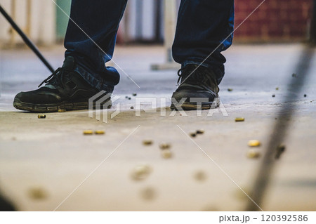 Close-up view of a person's feet. The person is wearing black shoes and dark pants. The ground appears to be covered with what looks like spent bullet casings or shells scattered around their feet 120392586