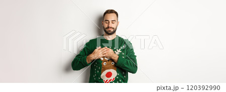 New year celebration and winter holidays concept. Young relaxed man feeling nostalgic, holding hands on heart and close eyes, dreaming about something on Christmas 120392990