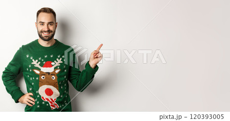 Winter holidays and christmas. Handsome bearded adult man pointing finger right at promo banner, standing in funny sweater against white background 120393005