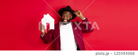 Real estate. Cheerful Black man showing paper house and smiling, recommending broker, standing over red background Real estate. Cheerful Black man showing paper house and smiling, recommending broker, standing over red background 120393008